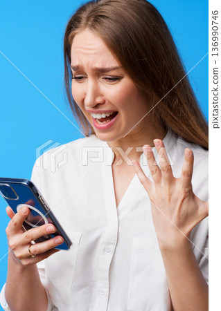 Woman reacts with surprise while looking at her phone on a bright blue background 136990746