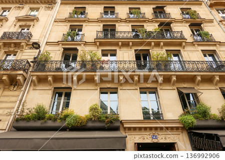 Paris classic haussmann building facade with ornate balconies, green plants, awnings, and large windows Paris classic haussmann building facade with ornate balconies, green plants, awnings, and large windows 136992906