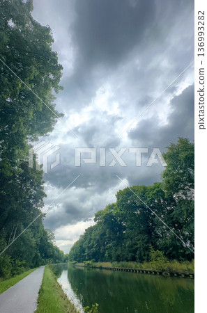 Tranquil pathway alongside reflective water beneath cloudy sky Tranquil pathway alongside reflective water beneath cloudy sky 136993282