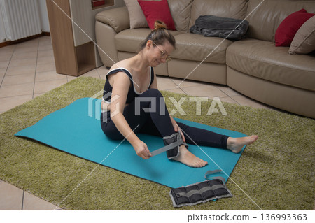 Woman sitting on an exercise mat, preparing for a home workout session and securing adjustable ankle weights to her leg, focusing on fitness and active lifestyle Woman sitting on an exercise mat, preparing for a home workout session and securing adjustable ankle weights to her leg, focusing on fitness and active lifestyle 136993363
