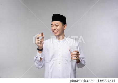 Young Asian individual excitedly anticipates Iftar with water and smile Young Asian individual excitedly anticipates Iftar with water and smile 136994245