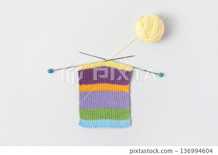 Striped piece of knitted fabric for future clothing with knitting needles and yellow ball of thread on gray background. Homemade needlework and knitting of warm, clothes and toys. Concept creativity Striped piece of knitted fabric for future clothing with knitting needles and yellow ball of thread on gray background. Homemade needlework and knitting of warm, clothes and toys. Concept creativity 136994604