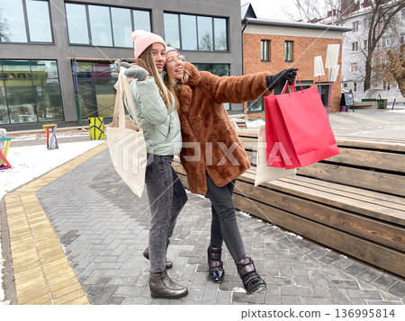 Two friends walking outdoors with shopping bags on a winter day. Shared excitement, fun, and urban lifestyle during cold weather and holiday mood. 136995814