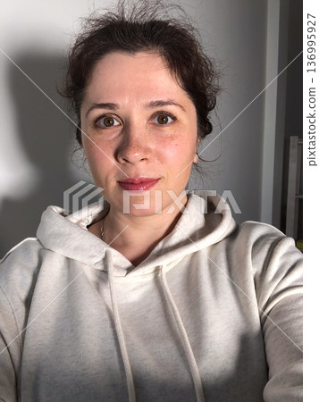 Selfie portrait of woman in cozy hoodie relaxing at home. Natural portrait in a modern domestic setting. Comfort in simplicity, everyday authenticity and serene domestic life concept 136995927