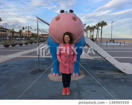 Young woman enjoying a delightful playful moment with a charming blue and pink hippo sculpture found in the playground Young woman enjoying a delightful playful moment with a charming blue and pink hippo sculpture found in the playground 136995930