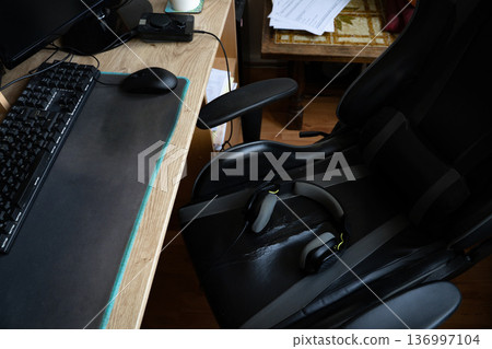 PC desk with gamer headphones resting on an old gaming chair in a casual home setup. Everyday real-life gaming scene showing workspace and tech gear. Concept of gaming, technology, setup, home PC desk with gamer headphones resting on an old gaming chair in a casual home setup. Everyday real-life gaming scene showing workspace and tech gear. Concept of gaming, technology, setup, home 136997104