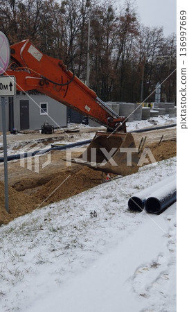 An orange excavator digs a dirt trench near black pipes in winter, construction site, industrial work. An orange excavator digs a dirt trench near black pipes in winter, construction site, industrial work. 136997669
