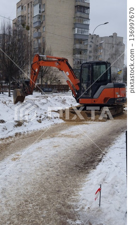 An orange excavator digs a trench in a snow-covered urban area near residential buildings, winter construction, heavy machinery. An orange excavator digs a trench in a snow-covered urban area near residential buildings, winter construction, heavy machinery. 136997670
