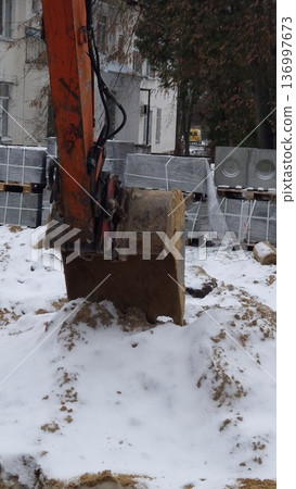 A close-up view of a dirty orange excavator bucket resting on a pile of snow and sand with building materials in the background, winter construction, heavy equipment. A close-up view of a dirty orange excavator bucket resting on a pile of snow and sand with building materials in the background, winter construction, heavy equipment. 136997673