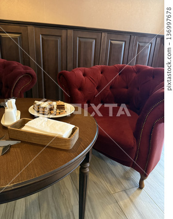 Small cakes and a napkin holder sit on a wooden table near red velvet armchairs, cozy cafe interior, relaxing lifestyle. Small cakes and a napkin holder sit on a wooden table near red velvet armchairs, cozy cafe interior, relaxing lifestyle. 136997678