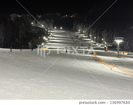 People skiing down an illuminated snowy slope at night, active winter lifestyle, extreme sports, winter resort. 136997680