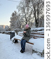 A smiling woman sits on a snowy park bench holding a small snowball, winter leisure, outdoor lifestyle, cold season fun. 136997693