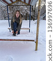 A woman sits on a snow-covered bench tying her shoelaces on a winter day, outdoor activity, cold season lifestyle, winter walk. 136997696