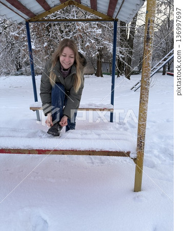 A smiling woman sits on a snowy bench tying her boots during a winter walk, outdoor lifestyle, cold season activity, winter leisure. A smiling woman sits on a snowy bench tying her boots during a winter walk, outdoor lifestyle, cold season activity, winter leisure. 136997697