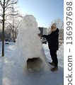 A woman stands next to a large snow fort with a tunnel entrance on a sunny winter day, outdoor recreation, creative winter fun, cold season activity. 136997698
