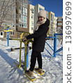 A smiling woman exercises on a yellow outdoor elliptical machine in a snowy courtyard on a bright winter day, active lifestyle, winter fitness, outdoor workout. 136997699