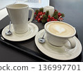 A black tray holds a white mug and a cappuccino with heart latte art next to a festive candle with red berries, cozy cafe moment, winter holiday spirit, Christmas. 136997701