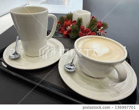 A black tray holds a white mug and a cappuccino with heart latte art next to a festive candle with red berries, cozy cafe moment, winter holiday spirit, Christmas. A black tray holds a white mug and a cappuccino with heart latte art next to a festive candle with red berries, cozy cafe moment, winter holiday spirit, Christmas. 136997701