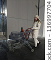 A woman pushes an empty shopping cart along a building on a sunny winter day, retail shopping, consumer lifestyle, outdoor errands. 136997704