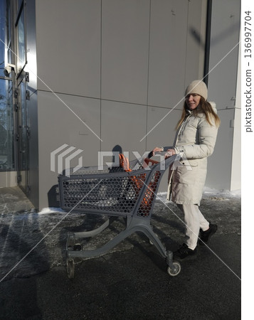 A woman pushes an empty shopping cart along a building on a sunny winter day, retail shopping, consumer lifestyle, outdoor errands. A woman pushes an empty shopping cart along a building on a sunny winter day, retail shopping, consumer lifestyle, outdoor errands. 136997704