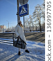 A woman in a winter coat stands by a snowy road holding a duffel bag and hailing a ride, winter travel, outdoor lifestyle, transportation concept. 136997705