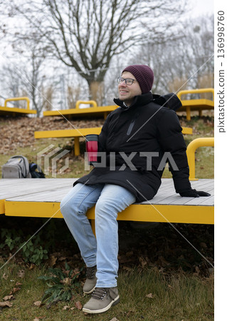 Young smiling man enjoying a warm drink from a red travel mug on a bank in winter. Concept of a city winter activity 136998760