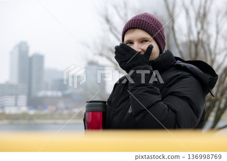 Young man enjoying a warm drink from a red travel mug near the river on a chilly day in winter. Concept of a city winter activity Young man enjoying a warm drink from a red travel mug near the river on a chilly day in winter. Concept of a city winter activity 136998769