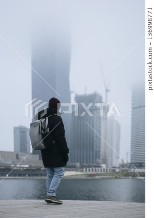 Man in winter coat overlooking a foggy city waterfront. Concept of a city winter activity Man in winter coat overlooking a foggy city waterfront. Concept of a city winter activity 136998771