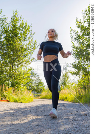 Woman running outdoors on a sunny trail surrounded by greenery and nature Woman running outdoors on a sunny trail surrounded by greenery and nature 136998948
