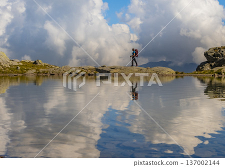 A hiker explores a serene alpine landscape in Swizz, surrounded by towering clouds and a calm lake. The reflection creates a picturesque scene of nature's beauty. A hiker explores a serene alpine landscape in Swizz, surrounded by towering clouds and a calm lake. The reflection creates a picturesque scene of nature's beauty. 137002144