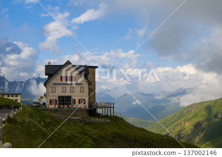 A house sits on a hillside in Switzerland, with mountains and clouds in the background. The area showcases natural beauty and outdoor scenery A house sits on a hillside in Switzerland, with mountains and clouds in the background. The area showcases natural beauty and outdoor scenery 137002146