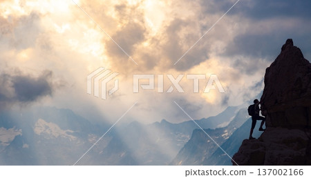A climber scales a rocky mountain peak as the sun sets, casting a golden glow over the landscape filled with distant peaks and clouds. A climber scales a rocky mountain peak as the sun sets, casting a golden glow over the landscape filled with distant peaks and clouds. 137002166