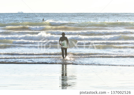 Surfer entering the ocean towards the evening sun Surfer entering the ocean towards the evening sun 137002526