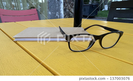 Glasses and notebook on a yellow table in an outdoor setting during daylight hours Glasses and notebook on a yellow table in an outdoor setting during daylight hours 137003076