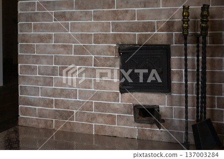 Brick fireplace with active fire. Fireplace tool set with ornate handles. Metal hearth protector. Warm inviting home interior Brick fireplace with active fire. Fireplace tool set with ornate handles. Metal hearth protector. Warm inviting home interior 137003284