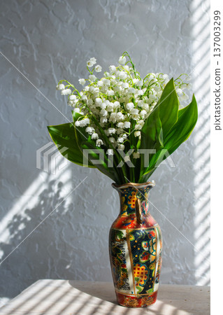 Spring flowers lily of the valley in decorative vase. Sunlight on windowsill. White bell-shaped blooms with large green leaves 137003299