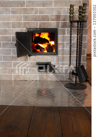 Fire burning in brick fireplace. Metal tools stand nearby. Traditional home heating with wood logs. Cozy rustic interior 137003302