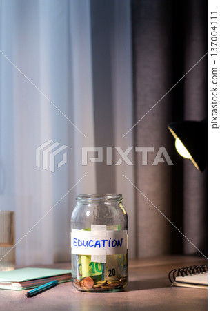 Hand dropping money into glass savings jar on table. Close up of fingers inserting cash into transparent container for education. Financial planning. University. Vertical Hand dropping money into glass savings jar on table. Close up of fingers inserting cash into transparent container for education. Financial planning. University. Vertical 137004111