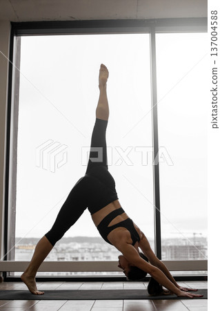 A woman in athletic wear performs a three-legged downward-facing dog yoga pose with one leg extended upwards, silhouetted against a large window overlooking a city skyline 137004588