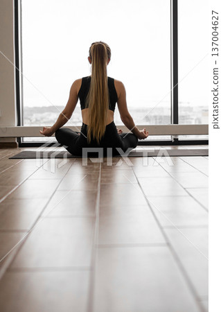 A woman with long blonde hair in a ponytail sits in a lotus pose on a yoga mat, facing a large window with an overcast sky visible outside A woman with long blonde hair in a ponytail sits in a lotus pose on a yoga mat, facing a large window with an overcast sky visible outside 137004627
