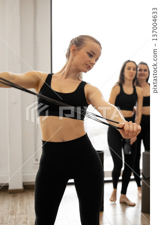 A woman in athletic wear stretches a resistance band while two other women stand behind her in a fitness studio A woman in athletic wear stretches a resistance band while two other women stand behind her in a fitness studio 137004633