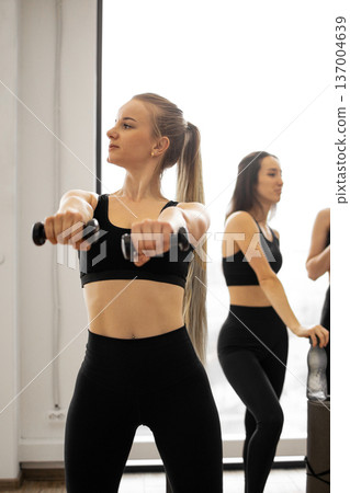 Three women in athletic wear are engaged in a fitness routine, holding dumbbells and focusing on their workout 137004639