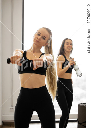 Two women in athletic wear are engaged in a fitness routine, one holding dumbbells and the other holding a water bottle 137004640
