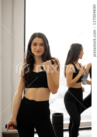 A woman in athletic wear holds dumbbells while looking at the camera, with other women exercising in the background A woman in athletic wear holds dumbbells while looking at the camera, with other women exercising in the background 137004690