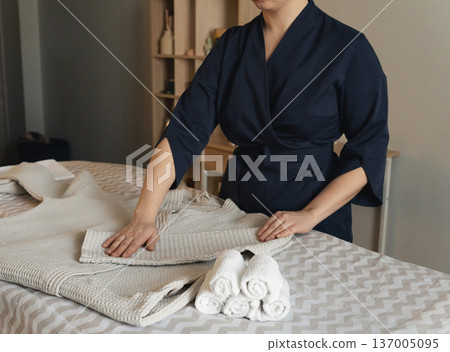 On massage table, a woman's hands fold a clean waffle robe next to neatly folded white towels. On massage table, a woman's hands fold a clean waffle robe next to neatly folded white towels. 137005095