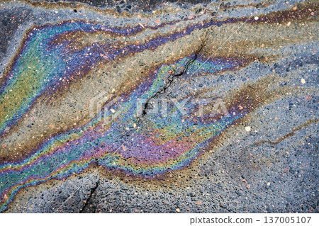Oil slick creating a vibrant iridescent rainbow pattern on cracked asphalt, depicting environmental pollution and abstract texture 137005107