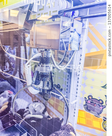 Close up of a claw machine grabber in a brightly lit arcade, attempting to pick up a stuffed toy prize from a pile. 137005614