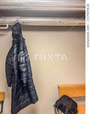 A long black winter down coat hangs from an industrial pipe in a simple, utilitarian locker room, a backpack on the bench. A long black winter down coat hangs from an industrial pipe in a simple, utilitarian locker room, a backpack on the bench. 137005616
