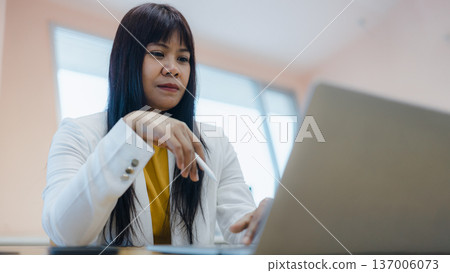 Focused Asian businesswoman using stylus pen with laptop in office. Professional female manager working on digital creative project or signing documents. Career success and modern technology. Focused Asian businesswoman using stylus pen with laptop in office. Professional female manager working on digital creative project or signing documents. Career success and modern technology. 137006073