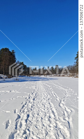 Winter landscape with snow covered frozen river and forest under clear blue sky with footprints on ice. Cold season and solitude concept Winter landscape with snow covered frozen river and forest under clear blue sky with footprints on ice. Cold season and solitude concept 137006722